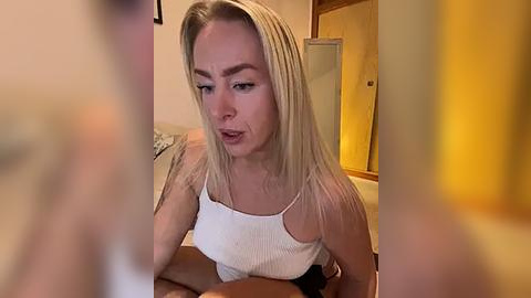 Media: Video of a blonde woman in a white tank top, sitting on a bed, with a blurry man in the foreground.
