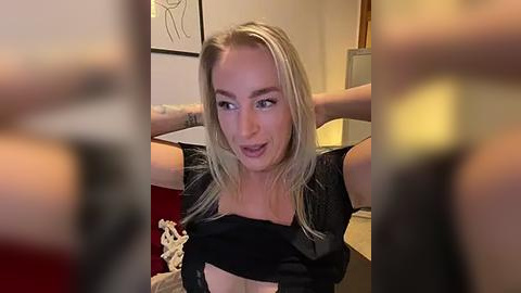 Media: Video of a blonde woman with fair skin, wearing a black top, stretching her arms behind her head, indoors, blurry background.