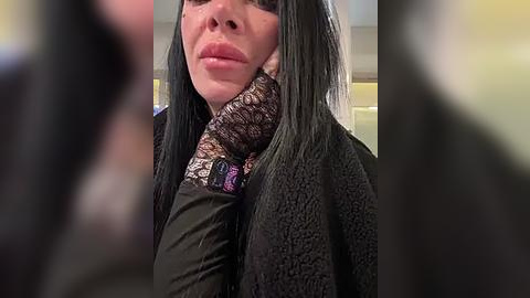 Media: A close-up video of a person with long black hair and fair skin, wearing a black textured coat with a patterned scarf. The background is blurred, with indistinct shapes and colors.