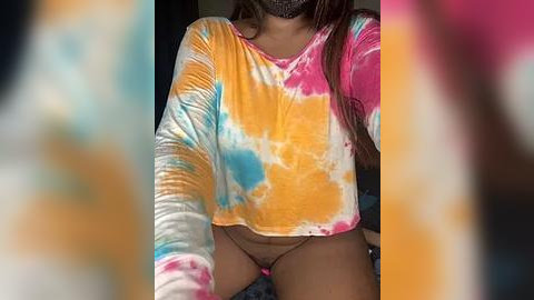 Media: Video of a young woman with medium brown skin, wearing a tie-dye crop top and black thong, with her long brown hair partially visible, seated on a bed, blurred background.