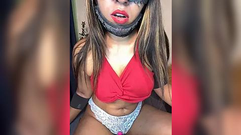 Media: Video of a young woman with medium-brown skin and long, straight hair, wearing a red halter top, lace panties, and a black blindfold. She sits on a bed, her legs spread.