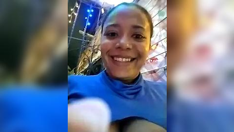 Media: Video of a smiling African-American woman with dark skin, medium build, and medium-length braided hair, wearing a blue turtleneck sweater. The background features a blurred, colorful, indoor setting with hanging decorations.