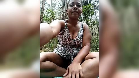 Media: Video of a smiling, dark-skinned woman with short hair, sitting cross-legged on a forest floor, wearing a floral-patterned top and shorts. Background shows blurred greenery.