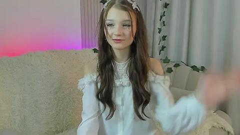 Media: Video of a young woman with long, wavy brown hair, wearing a white, off-the-shoulder lace dress with a pearl necklace, sitting on a cream-colored couch in a softly lit room with purple and pink ambient lighting.