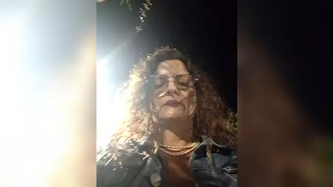 Media: A video of a middle-aged woman with curly hair, glasses, and a denim jacket, captured in a dimly lit, outdoor setting with lens flare.