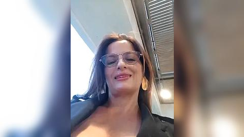 Media: Video of a middle-aged woman with medium skin tone and long brown hair, wearing clear glasses and a black leather jacket, smiling, taken from a low angle, indoors near a window with blinds.