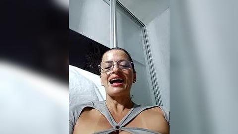 Media: Video of a smiling woman with fair skin, brown hair tied back, wearing glasses and a gray, cut-out top. She stands in a modern bathroom with a glass shower enclosure in the background.