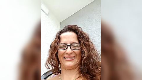 Media: A video of a woman with curly brown hair and glasses, smiling, standing against a white wall. She is wearing a necklace.