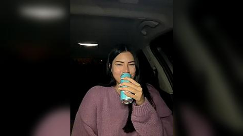 Media: A video of a young woman with long black hair, wearing a lavender sweater, drinking from a blue can, inside a dimly lit car at night.