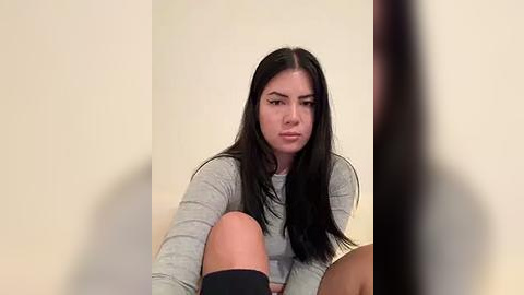 Media: Video of a young woman with long black hair, wearing a gray sweater, sitting on a bed with a beige wall background.