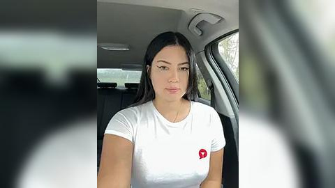 Media: Video of a young woman with long dark hair, wearing a white t-shirt with a red logo, seated in a car, looking directly at the camera.