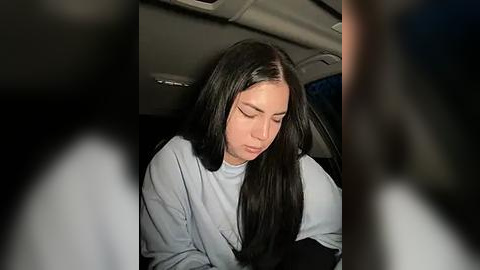 Media: Video of a young woman with long, straight black hair, wearing a light blue sweater, sitting in a car, eyes closed, appearing tired.