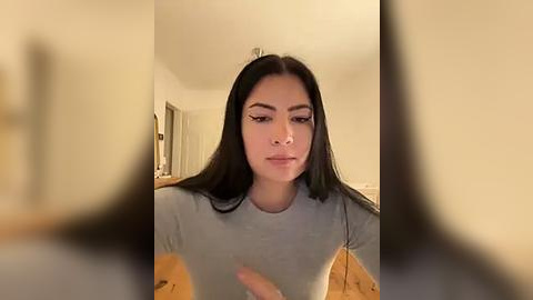 Media: Video of a young woman with long, straight black hair and fair skin, wearing a light gray top, sitting in a dimly lit, cozy room with beige walls and wooden floors.