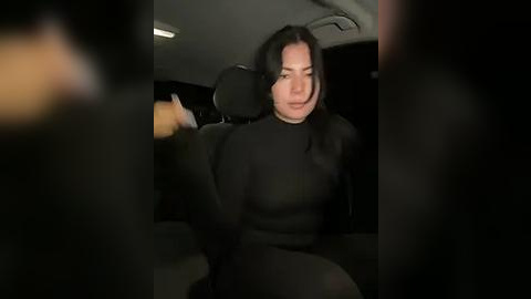 Media: Video of a woman with long black hair, wearing a black sweater, sitting in a car. Her expression is neutral. The background shows a dark interior.