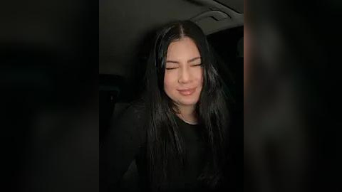 Media: Video of a woman with long black hair, wearing a black top, smiling gently in a dimly lit car interior.