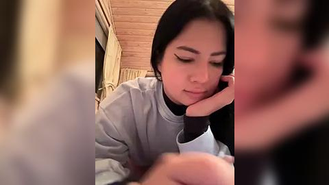 Media: Video of a young woman with long black hair, wearing a light gray hoodie, resting her head on her hand, indoors with wooden walls and ceiling.