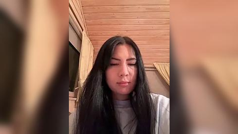 Media: Video of a young Asian woman with long black hair, wearing a grey top, standing in a wooden-paneled room with beige curtains and a window. She appears to be in a contemplative or reflective state, eyes closed, with a serene expression.