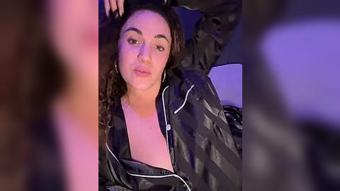 Media: Video of a young woman with medium skin tone and curly brown hair, wearing a black leather jacket, lying on a couch with a purple light.