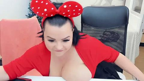 Media: Video of a fair-skinned woman with dark hair, wearing a red top and large red polka dot Minnie Mouse ears, sitting at a desk with a laptop. Background shows office furniture and plants.