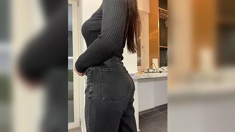 Media: Video of a woman in a black ribbed sweater and high-waisted, dark denim jeans, standing with hands on hips in a modern kitchen with white cabinets, a sink, and a tiled floor.