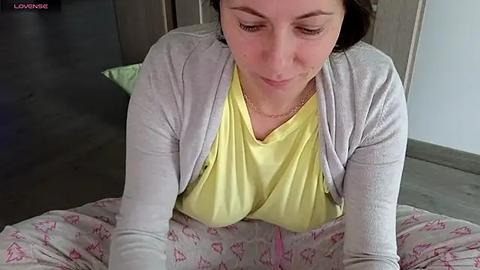 Media: Video of a light-skinned woman with shoulder-length brown hair, wearing a yellow t-shirt and light gray cardigan, lying on a bed with pink patterned sheets, eyes closed, in a cozy bedroom.