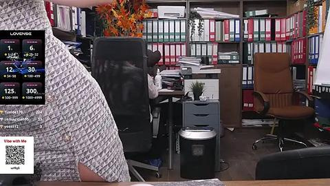 Media: Video of a messy office with a person in a white patterned shirt, a woman sitting at a desk, and colorful bookshelves in the background.