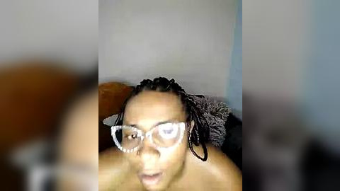 Media: Video of a shirtless Black man with dreadlocks and white-framed glasses, sitting on a couch with a brown cushion. Background is blurred, possibly indoors.