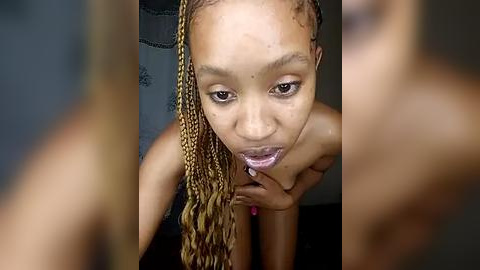 Media: Video of a topless Black woman with medium-brown skin and long, braided hair, licking her lips while gazing upward. The background is blurred, focusing on her facial expression.