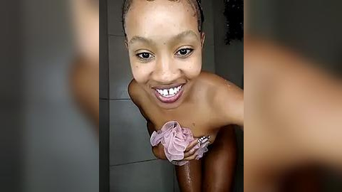 Media: Video of a smiling, light-skinned Black woman with short hair in a shower, holding a pink loofah. Water droplets glisten on her skin.