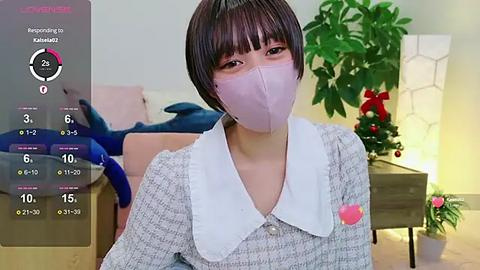 Media: A video shows a young Asian woman with short black hair, wearing a light pink face mask, gray textured blouse, and a white collar, sitting in a cozy living room with Christmas decorations.