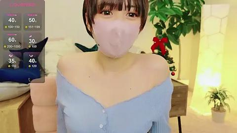 Media: Video of a young Asian woman with short brown hair, wearing a light blue off-shoulder top and a pink face mask, in a cozy, lit room with a Christmas tree and potted plants.