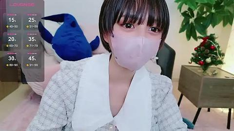 Media: Video of a young East Asian woman with short black hair, wearing a white mask, white blouse, and gray patterned cardigan, sitting in a room with a blue stuffed shark toy, a Christmas tree, and a wooden table.