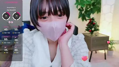 Media: A video of an Asian woman with short black hair, wearing a face mask, white bathrobe, and seated indoors, next to a decorated Christmas tree.