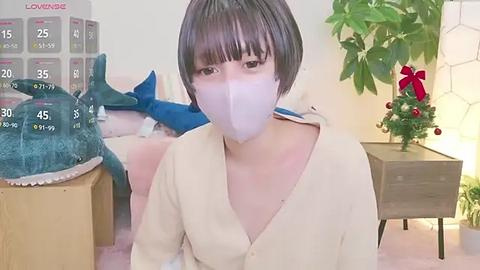 Media: A video of an Asian woman with short black hair and pale skin, wearing a white mask, beige cardigan, and sitting in a cozy room with a Christmas tree, calendar, and a shark toy.