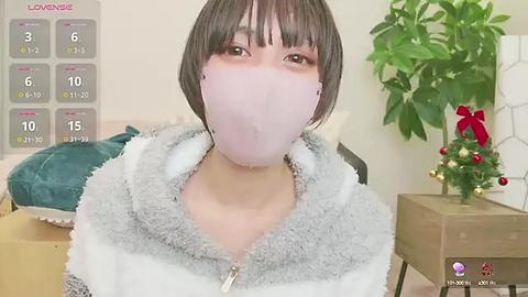 Media: A video of a young Asian woman with short, dark hair, wearing a light pink mask and a gray and white fuzzy hoodie, sitting in a room with a calendar, plant, and Christmas decorations.