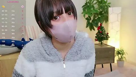 Media: Video of an Asian woman with short black hair and a white face mask, wearing a gray and white striped hoodie, indoors with a potted plant, a wall-mounted clock, and a lamp.