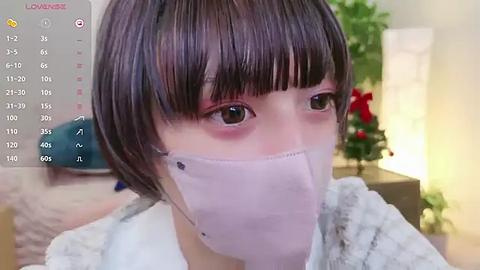 Media: A video of a young Asian woman with short, dark hair and pink eye shadow, wearing a white surgical mask and a light gray sweater, indoors with a blurred background.