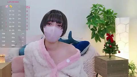 Media: A video shows an Asian woman with a short bob haircut and fair skin wearing a pink face mask and fluffy white bathrobe. She stands in a modern room with a potted plant, Christmas tree, and a calendar.