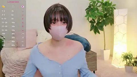 Media: A video of an Asian woman with a bob haircut, wearing a light blue off-shoulder blouse, a face mask, and sitting in a cozy, minimalist room with a green plant and a hexagonal lamp in the background.