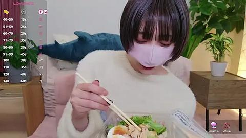 Media: Video of an Asian woman with short black hair, wearing a white mask and fluffy sweater, eating ramen with chopsticks on a couch.