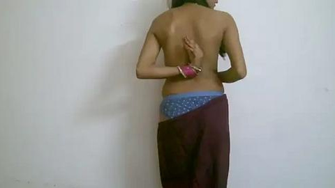 Media: Video of a topless South Asian woman with medium brown skin, wearing blue polka-dot panties and a maroon skirt. She stands against a plain white wall, with a pink bracelet on her right wrist.