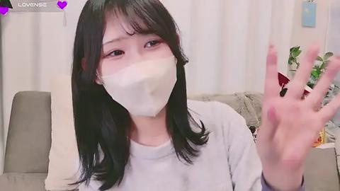 Media: A video of an Asian woman with long black hair and fair skin, wearing a white mask and a gray shirt, waving with a pink-painted fingernail, indoors with beige curtains and a green chair.