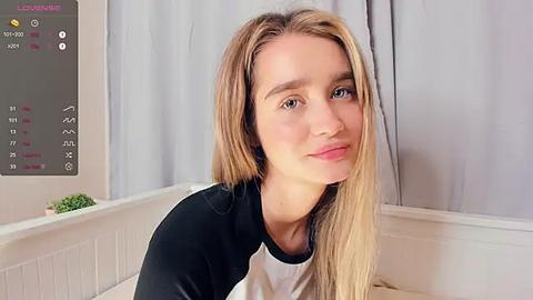 Media: Video of a young Caucasian woman with long blonde hair, fair skin, and blue eyes, wearing a black and white shirt, sitting indoors with a white radiator and potted plant in the background.