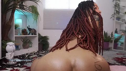 Media: Video of a nude woman with dark brown skin and long, maroon dreadlocks, sitting on a patterned bed, in a room with potted plants, a white shelf, and a window.