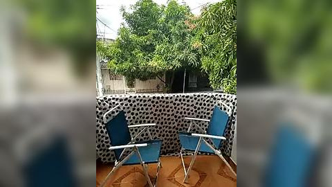 Media: Video of a small, cozy outdoor patio with two blue folding chairs and a brown table. The background features a white wall with black polka dots and lush green trees, creating a vibrant, inviting setting.