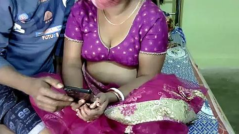 Media: Video of an Indian woman in a purple saree, with a man in a blue shirt and shorts, holding a phone, in a simple indoor setting.