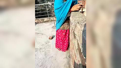 Media: Video of a person standing with a bright blue shawl and a red patterned cloth, against a rustic, beige wall with a metal railing and a sandy ground.