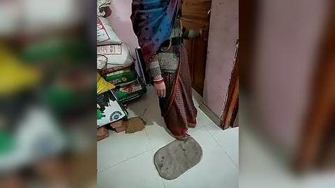 Media: Video of a person wearing a tie-dye shirt and brown pants standing on a cracked tile floor with a hole. Background shows stacked bags, a wooden door, and a blurred figure.