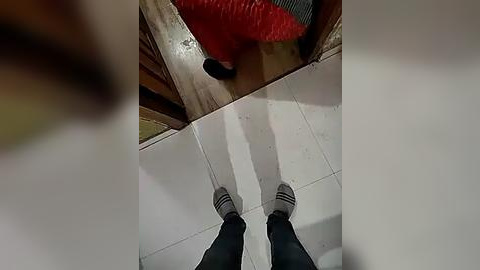 Media: A video of a person's feet in black socks and gray shoes standing on a tiled floor. The background includes a red, textured cushion and a wooden surface. The image is taken from a low-angle perspective.