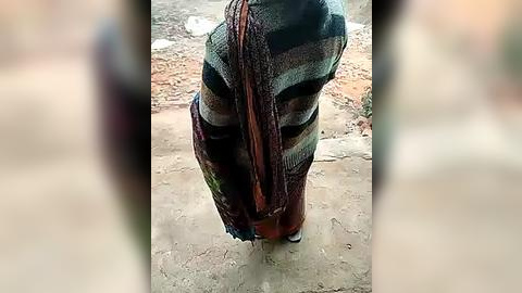 Media: A video of a person from the waist down, wearing a colorful, striped shawl and brown pants, standing on a dirt ground with a blurred background.